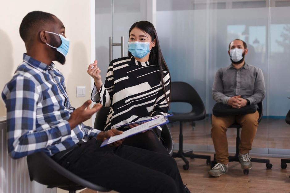 A patient discussing with a Cigna Insurance Psychiatrist in Soldiers Field Court in a group session, holding a clipboard, with others observing in a modern office setting