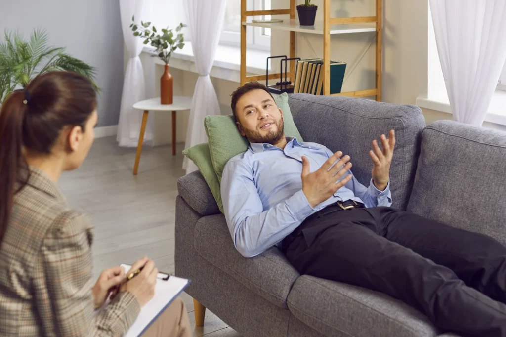 Man on therapy couch gesturing to a United Healthcare psychiatrist in Sugar Land, in a modern office with plants and books for effective mental health support.