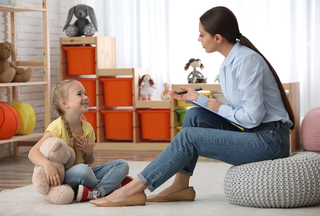 Smiling girl sharing thoughts with counselor holding clipboard amid toys in a playroom for same-day ADHD testing in Sugar Land, promoting engaging and supportive evaluation experiences.