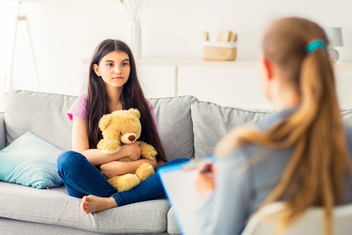 Adolescent Depression Psychiatrist Sugar Land counseling session: teen hugging teddy on couch during therapy in modern office, supportive care.