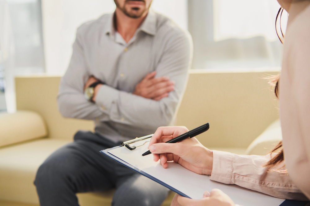 Psychiatrist taking notes while listening to male patient in therapy office, providing professional care through United Healthcare Psychiatrist Sugar Land services.