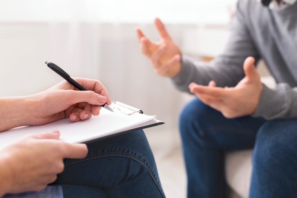 Close-up of an adhd doctor Sugar Land writing on a clipboard while a patient in a grey sweater gestures in talk therapy.