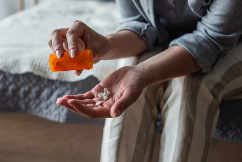 A patient pours prescribed depression medication Sugar Land pills from an orange bottle into their hand while sitting on a bed at home.