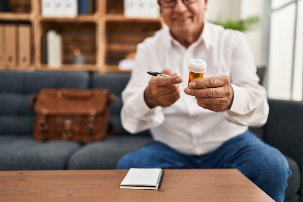 A senior doctor holds a pill bottle and points with a pen, explaining the proper dosage of depression medication Sugar Land to a patient.