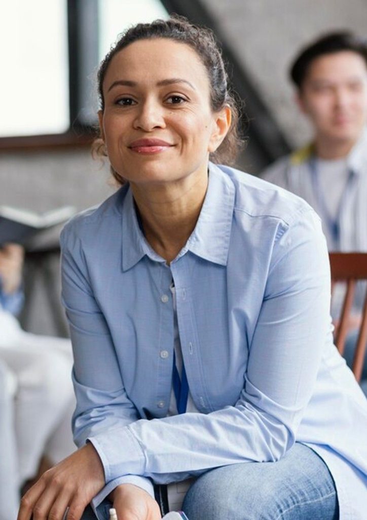 A smiling woman in a light blue button-down shirt sitting in a bright office, representing the positive outcomes of visiting a trusted psychiatrist in Sugar Land.