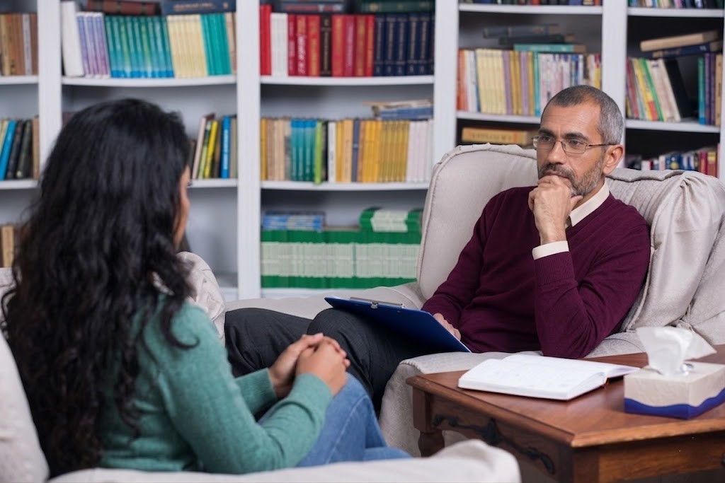 A male Anthem insurance psychiatrist in Sugar Land listens to a woman's concerns during a therapy session in a library.